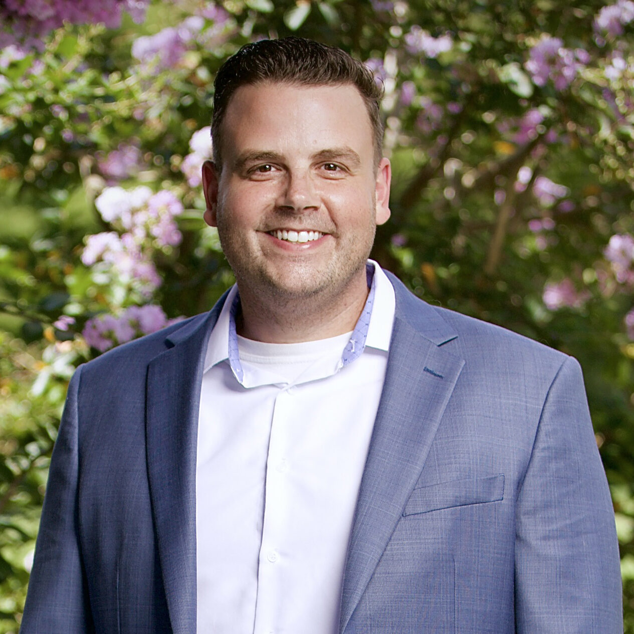 A smiling man in a blue blazer stands outdoors with flowering trees in the background.